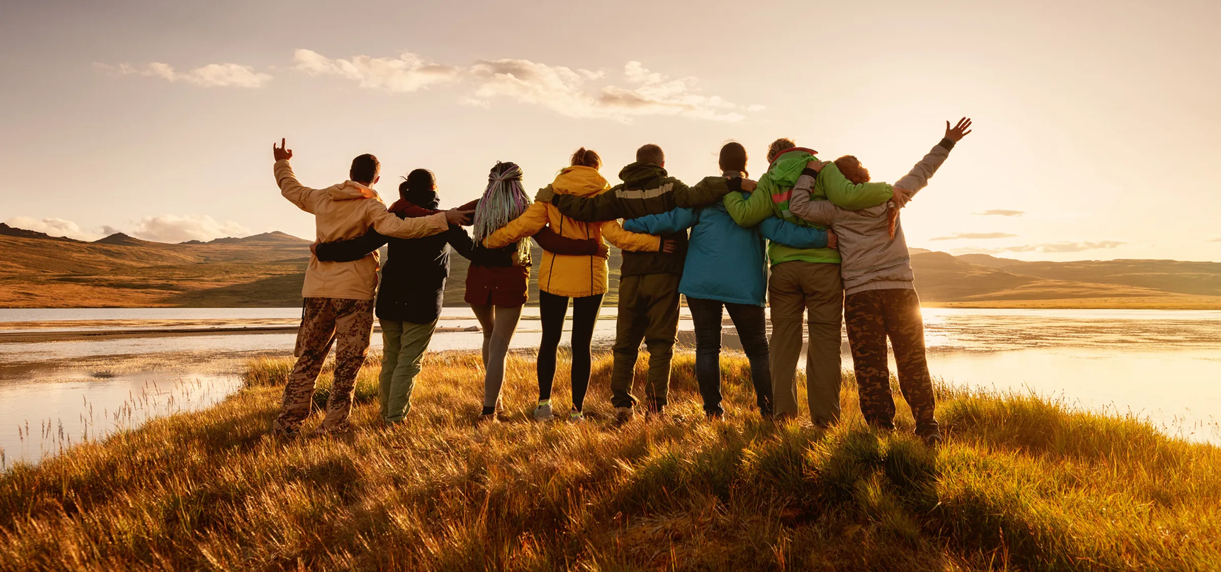 As the sun rises over a body of water, a group of people stand watching on a grassy field with arms wrapped around each other’s backs.